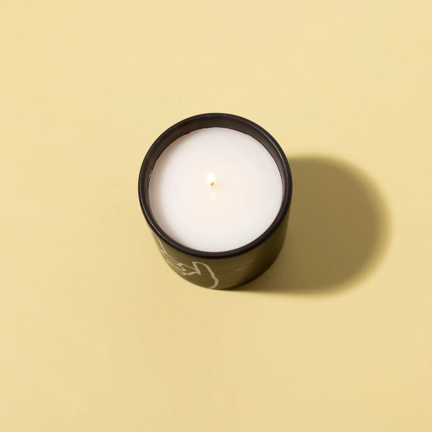 The Impressions 5.75 oz Candle - Leather + Oakmoss Rock On features a lit white matte ceramic candle in a black container, placed on a light yellow surface and casting a gentle shadow to the right.