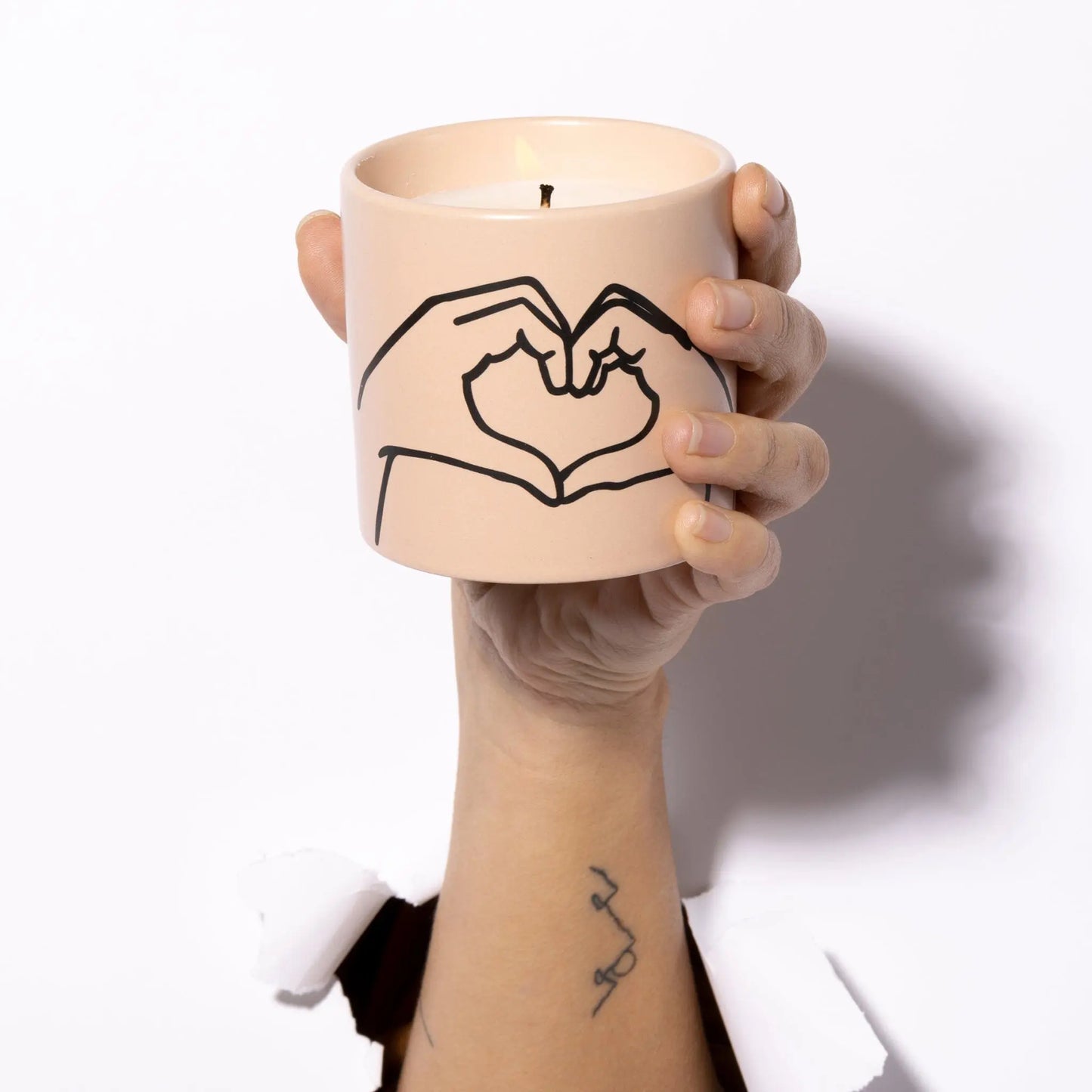 A hand holds the Impressions 5.75 oz Candle - Tobacco + Vanilla Love Ya, featuring a simple line drawing of hands forming a heart; part of an arm and tattoo peek through a torn white background.