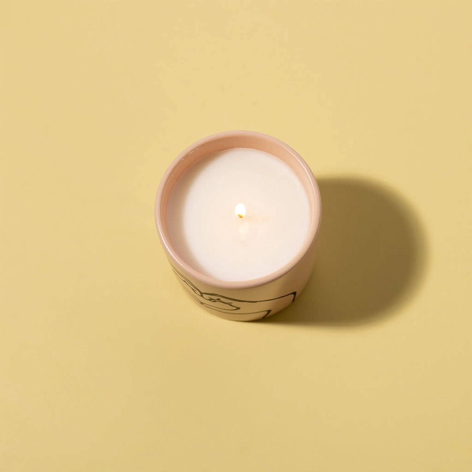 The Impressions 5.75 oz Love Ya Candle in Tobacco + Vanilla sits in a light pink matte ceramic holder on a pale yellow surface, casting a gentle shadow to the right.