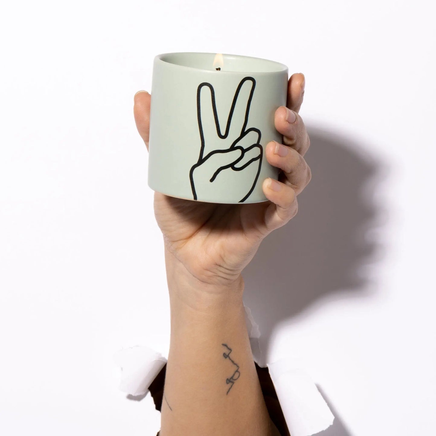 A hand breaks through white paper holding the Impressions 5.75 oz Candle - Lavender + Thyme Peace, a light green candle with a black peace sign outline; it’s lit and delicately scented with crushed lavender.