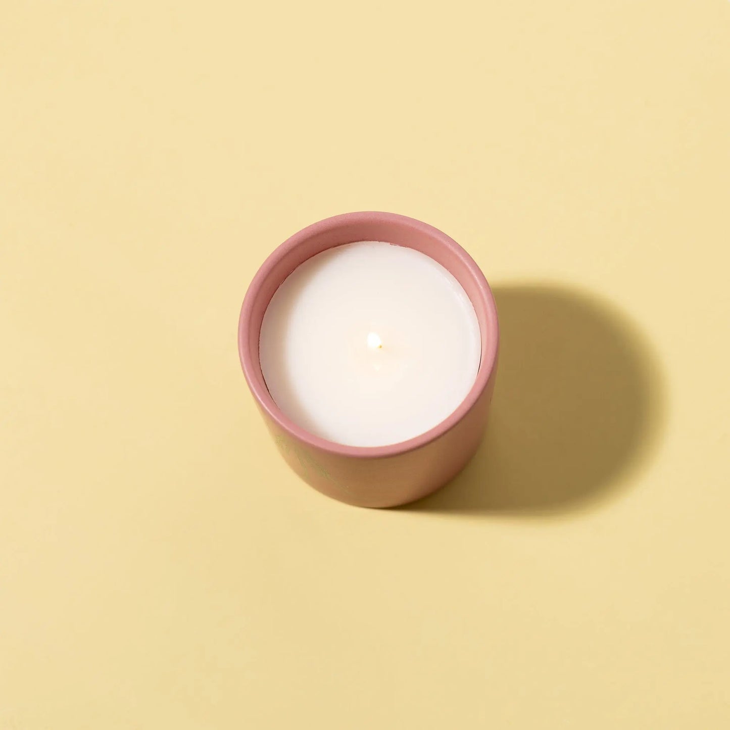 The Impressions 5.75 oz Candle - Violet Vanilla Thankful For You, in a pink matte ceramic holder with white wax, sits on a pale yellow surface and casts a shadow to the right.