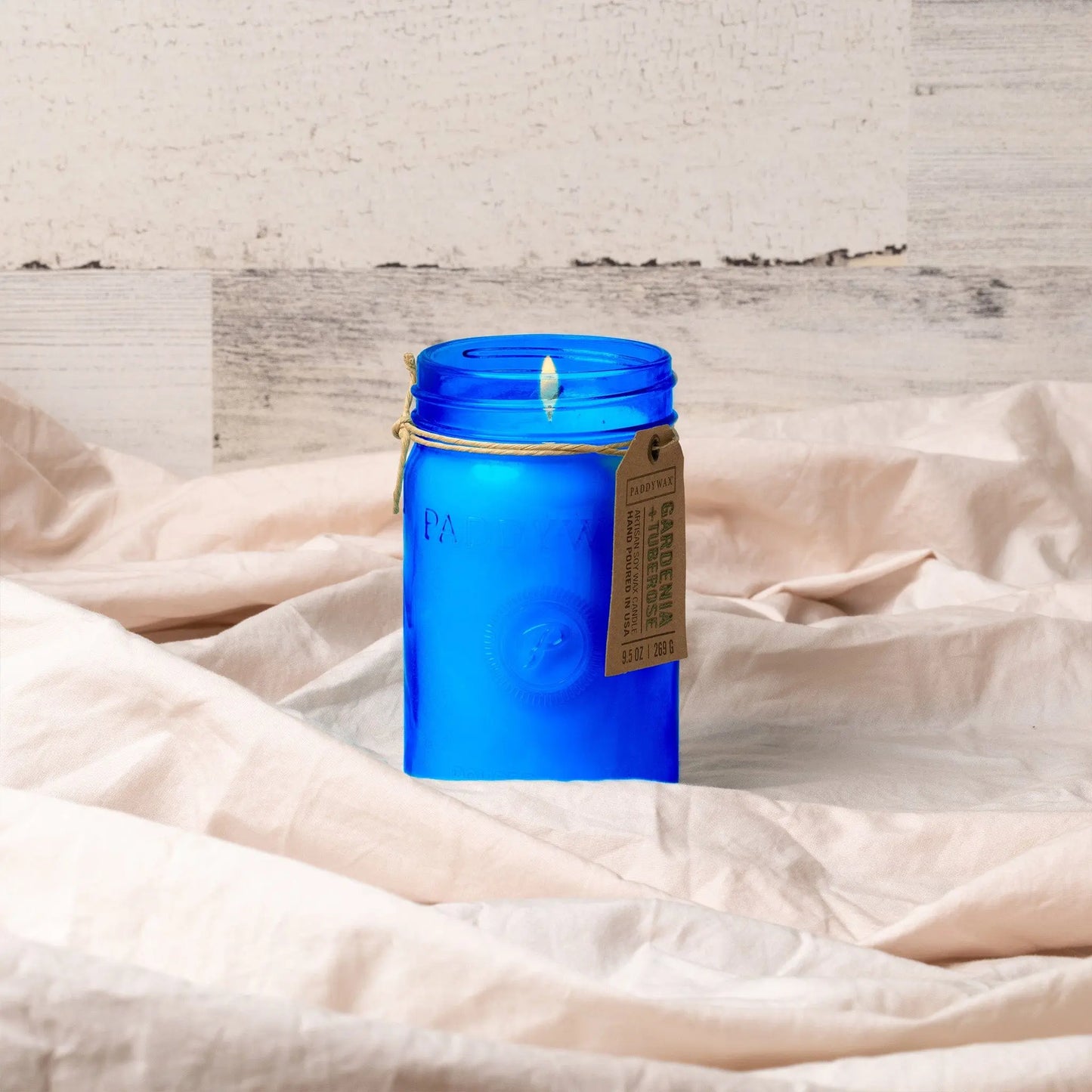 The Relish 9.5 oz Candle - Gardenia & Tuberose, in a blue glass jar with a brown string tag, sits on light fabric as its calming scent fills the air—an inviting piece from the Relish collection.
