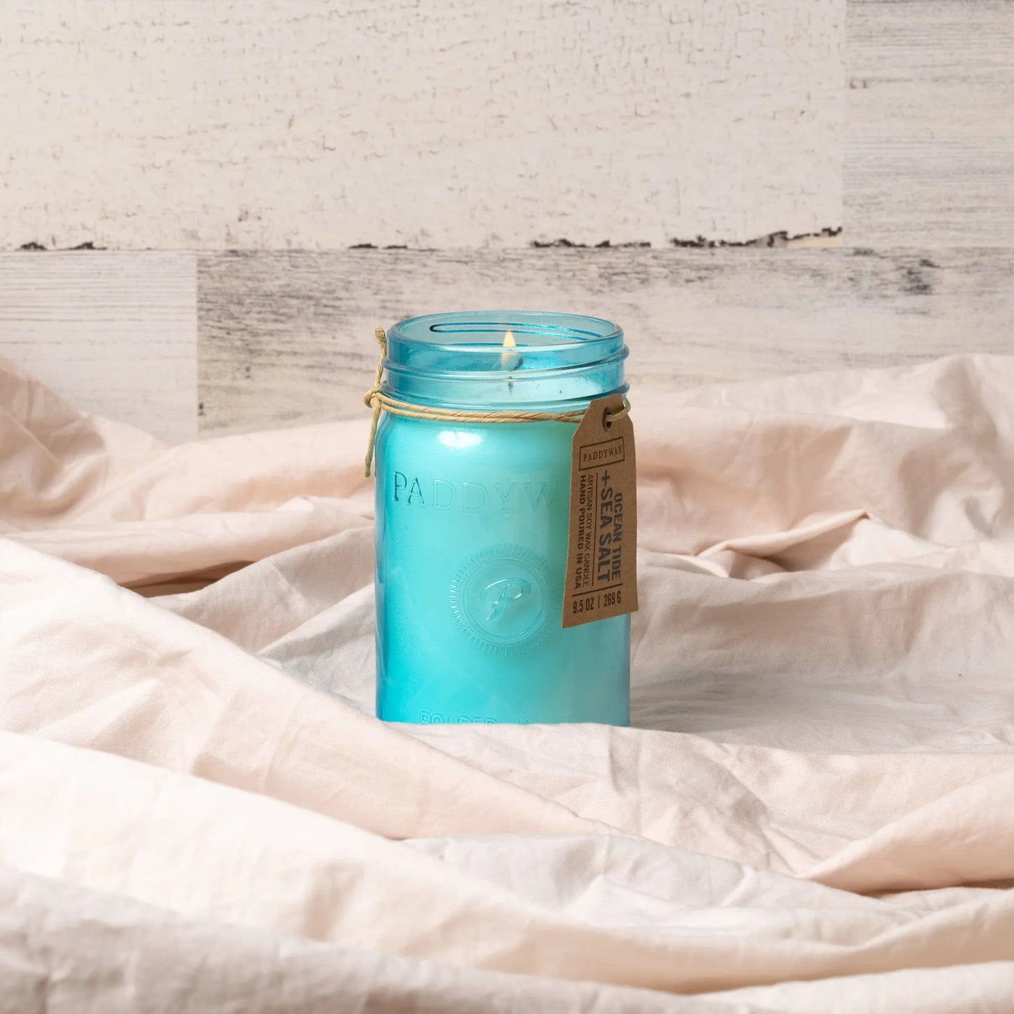 The Relish 9.5 oz Candle - Ocean Tide & Sea Salt, in a blue glass jar with a brown tag and lit wick, adds fragrance and style to your space on beige fabric with a light wood background.