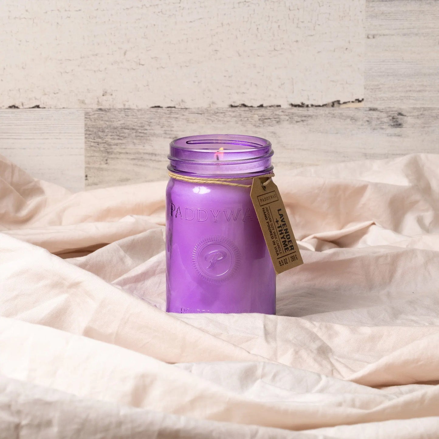 A Relish 9.5 oz Candle - Lavender + Thyme sits on beige fabric with a light wooden background, featuring its signature purple glass jar and offering a soothing fragrance.