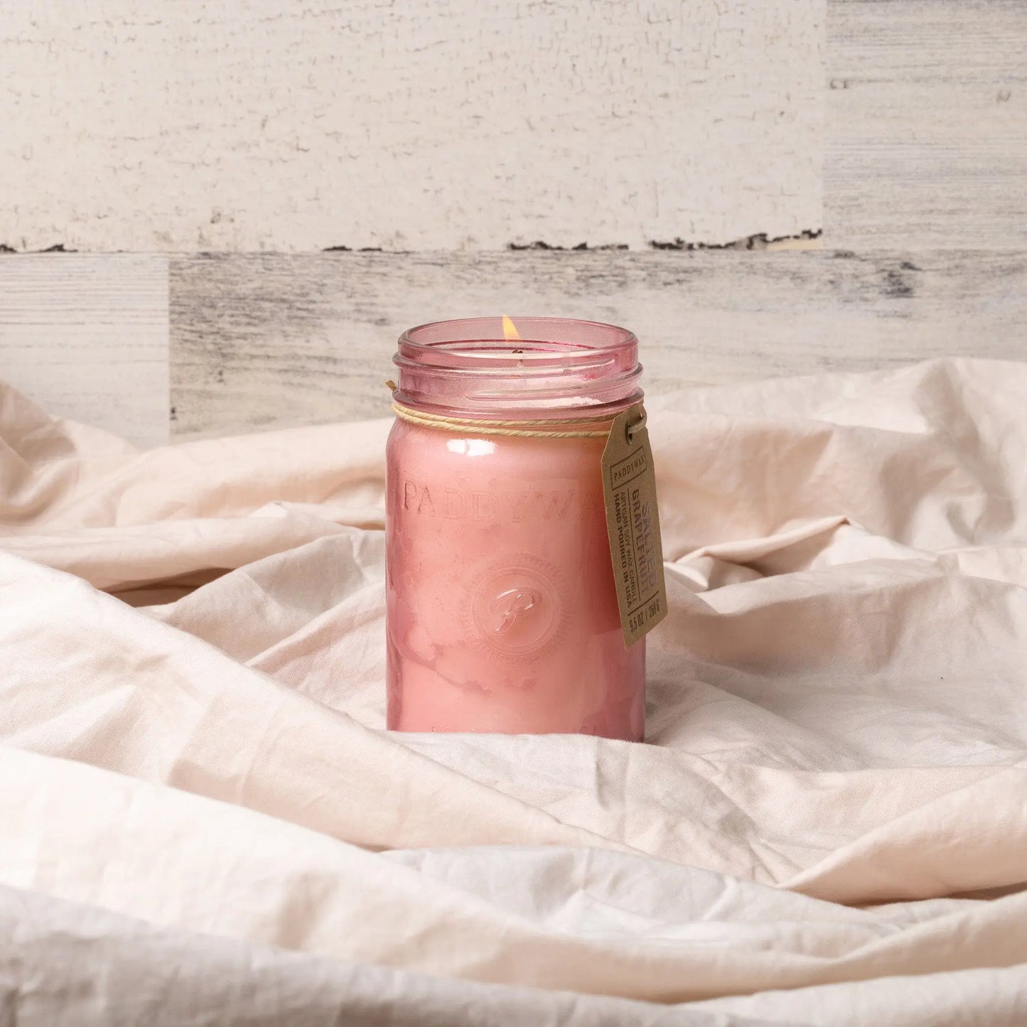 A lit Relish 9.5 oz Candle - Salted Grapefruit glows in its fragrant jar, resting on soft beige fabric with a textured light wall behind it.