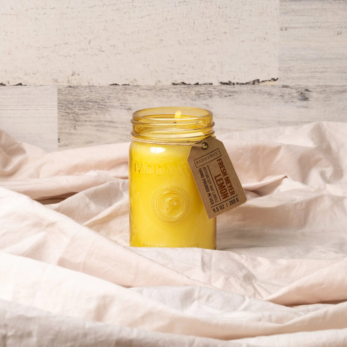 The Relish 9.5 oz. Candle - Fresh Meyer Lemon adds fragrance to any space, featuring a yellow candle in a glass jar with a tag, displayed on light, wrinkled fabric against a neutral background.