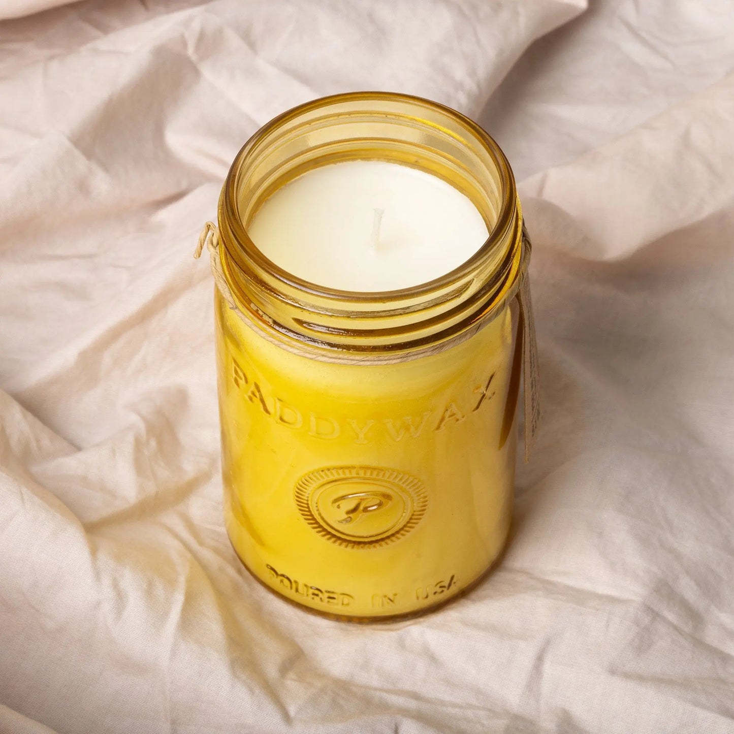 A Relish 9.5 oz. Candle in Fresh Meyer Lemon sits on a wrinkled light fabric, its yellow glass jar adding a bright fragrance to your space.