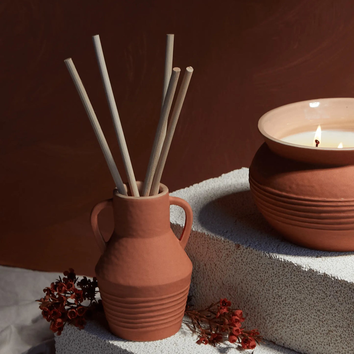 A Santorini 4 fl oz. Diffuser in Raw Clay & Pear, featuring a terracotta vase with reed sticks and red flowers, sits beside a lit candle on a textured white surface.