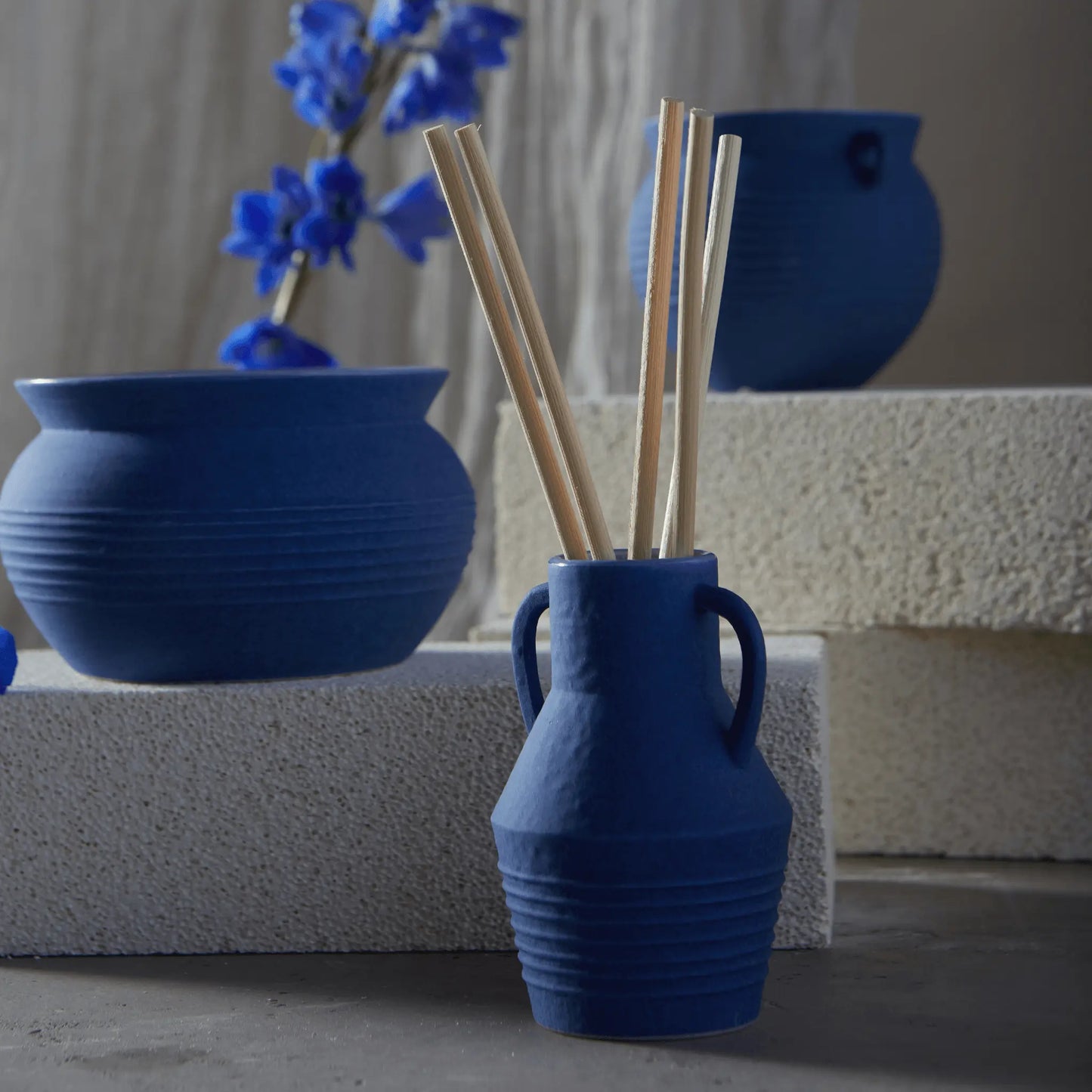 The Santorini 4 fl oz. Diffuser - Salted Blue Agave and coordinated blue ceramic vases are arranged on light textured blocks, with the diffuser holding wooden sticks and blue flowers in the background.