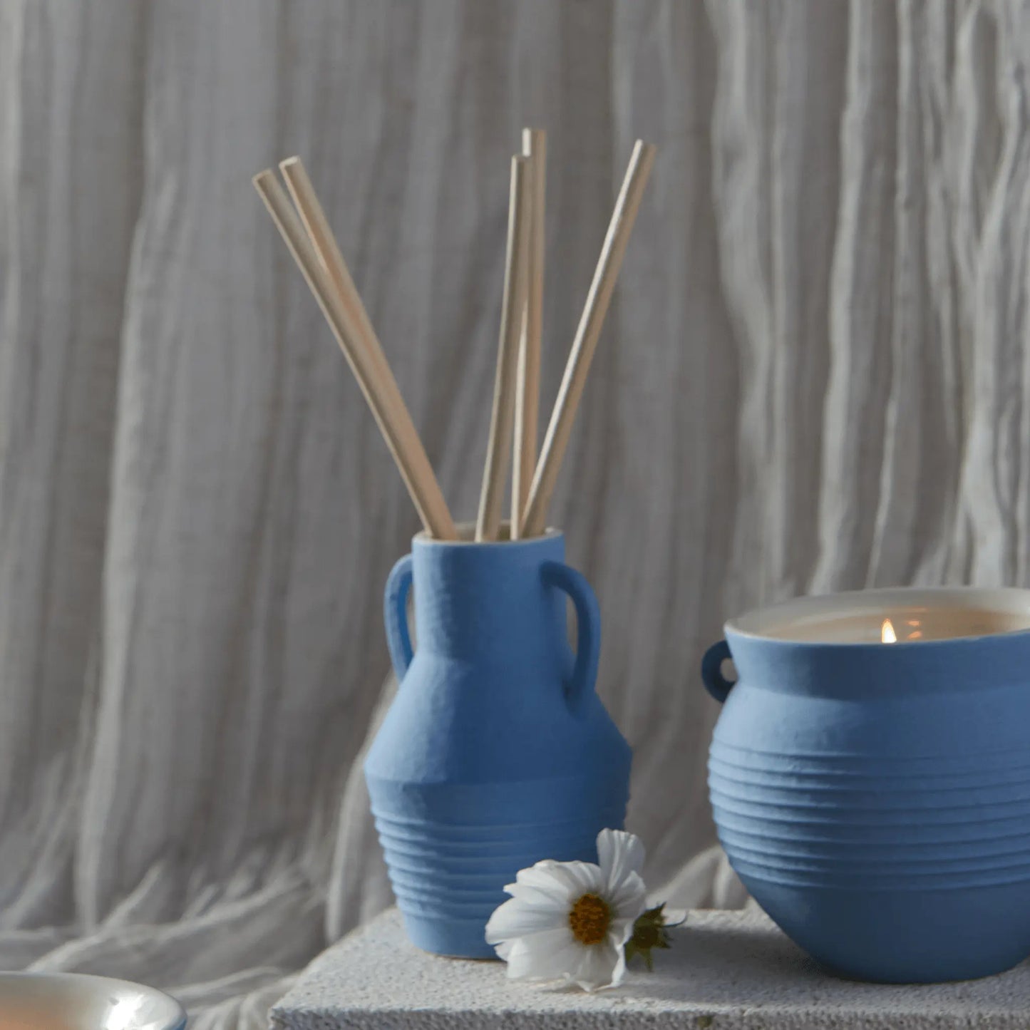 The Santorini 4 fl oz. Diffuser - Rosemary Sea Salt holds wooden sticks and is styled next to a blue candle jar and a white daisy, all set against a textured neutral background.