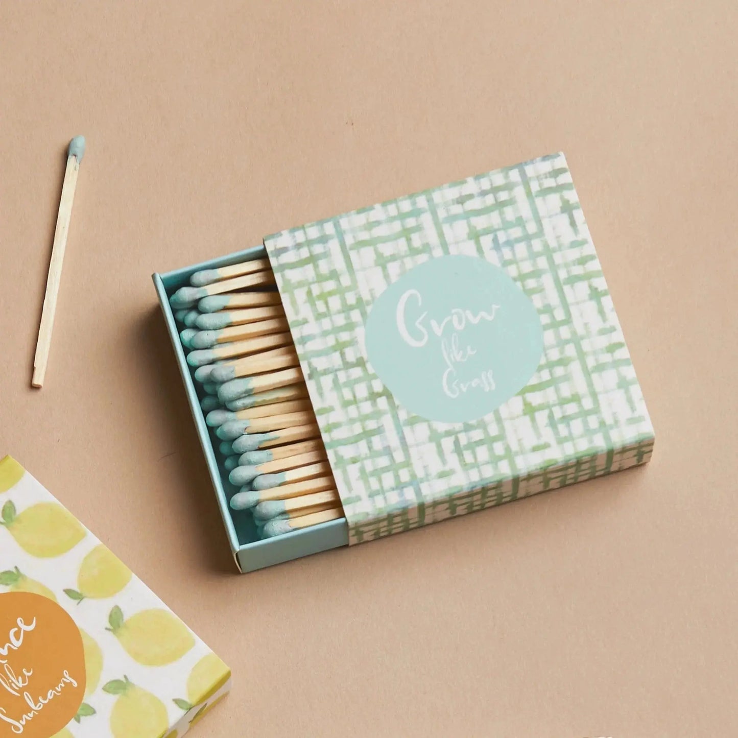 An open box of Terrace Matches - Grow Like Grass with blue-tipped safety matches rests on a beige surface. The green and white box displays the message Grow like Grass, making it an ideal complement to any candle collection.