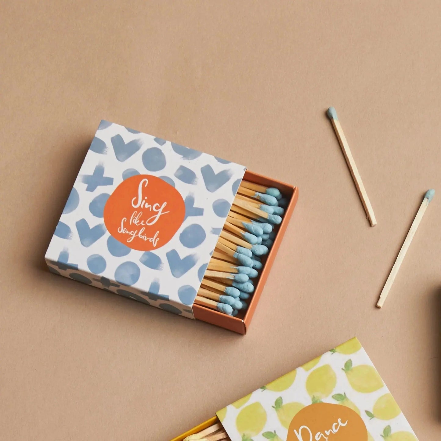 A box of Terrace Matches - Sing Like Songbirds, featuring a patterned cover and blue-tipped matches, rests on a beige surface with some matches scattered nearby—a delightful accent for any candle collection.