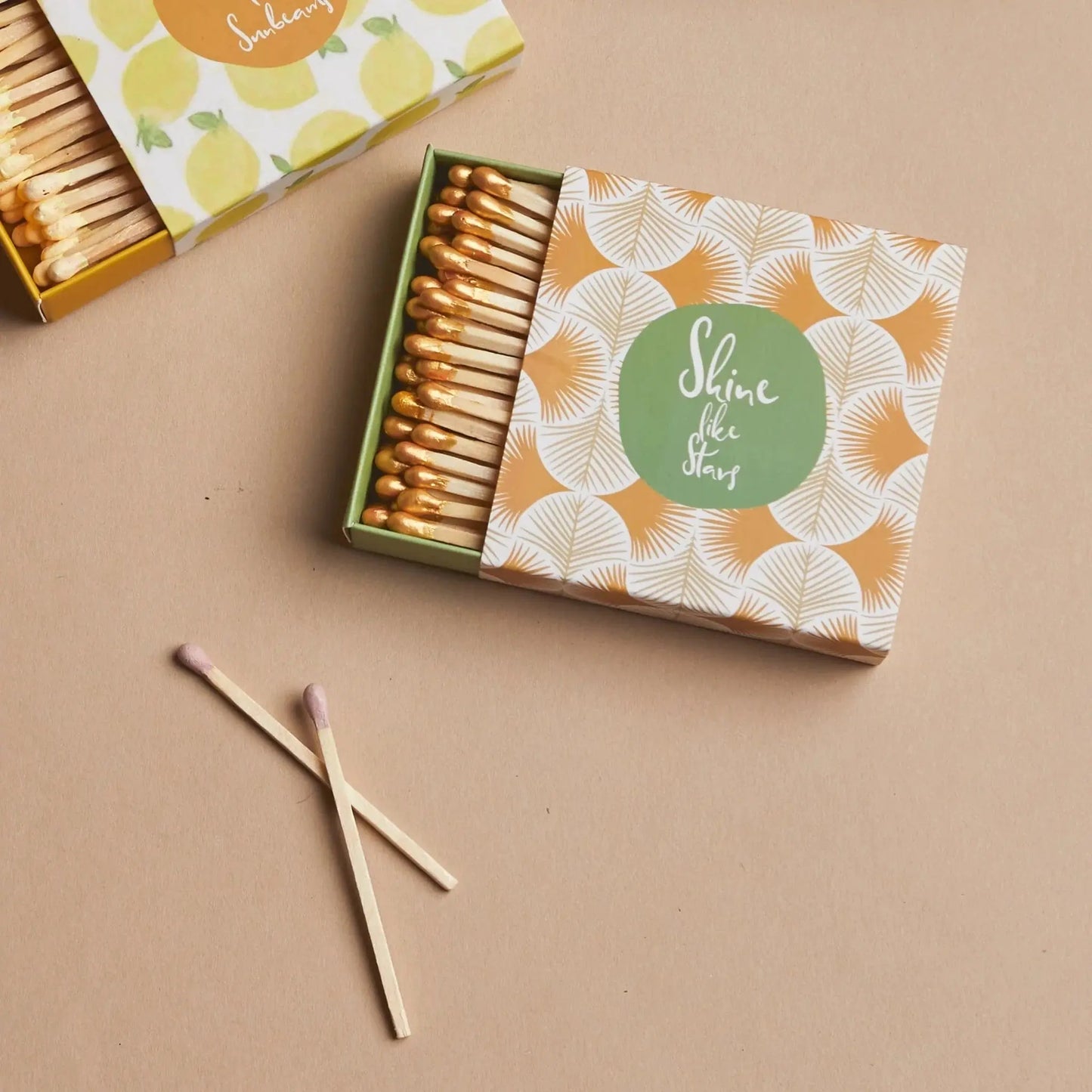 An open box of Terrace Matches - Shine Like Stars with a geometric design sits on a beige surface beside two used matches, making this stylish set a charming gift that pairs perfectly with any candle collection.