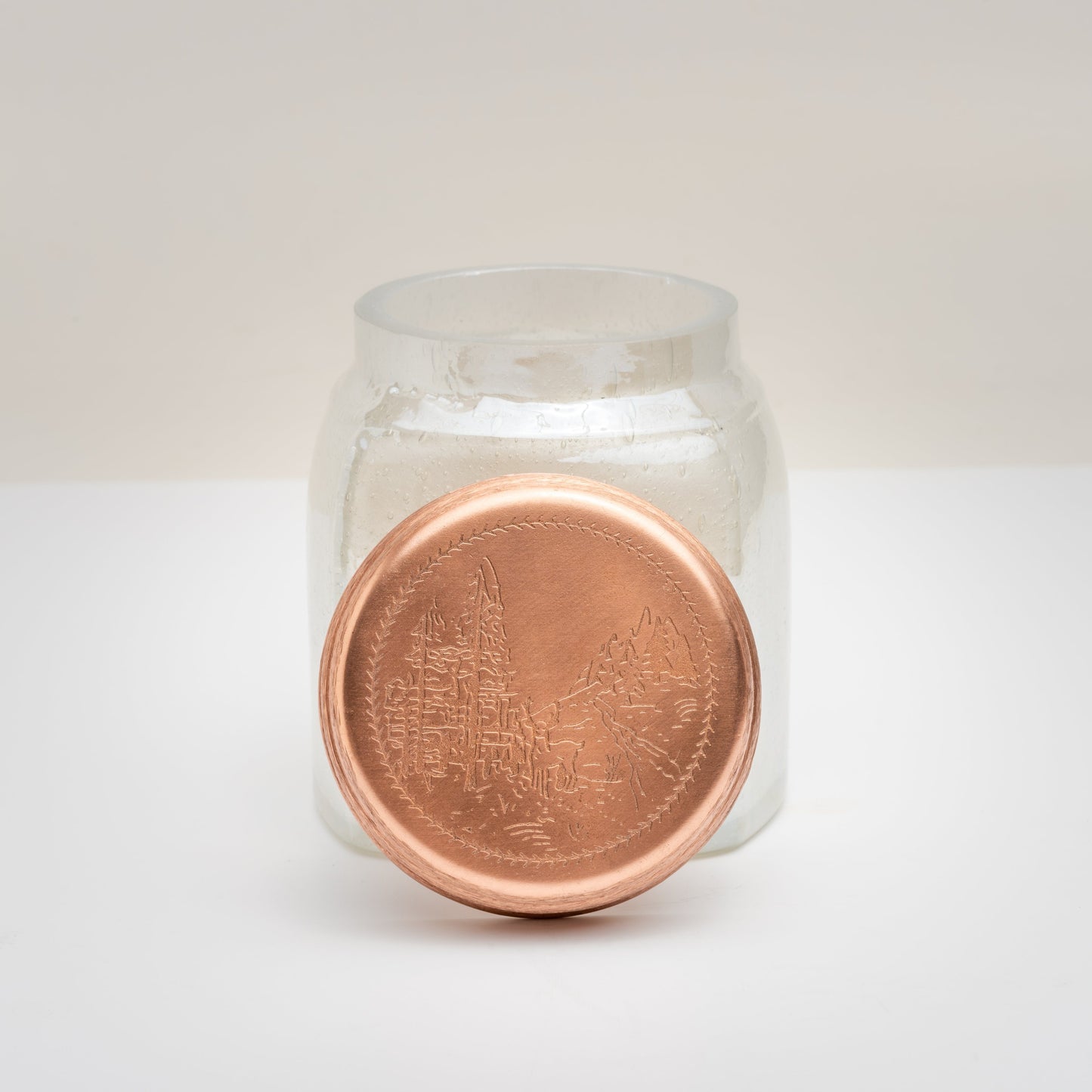 14 oz. Cypress & Fir Bubble Glass Candle with lid taken off; copper lid features an etched landscape full of fir trees