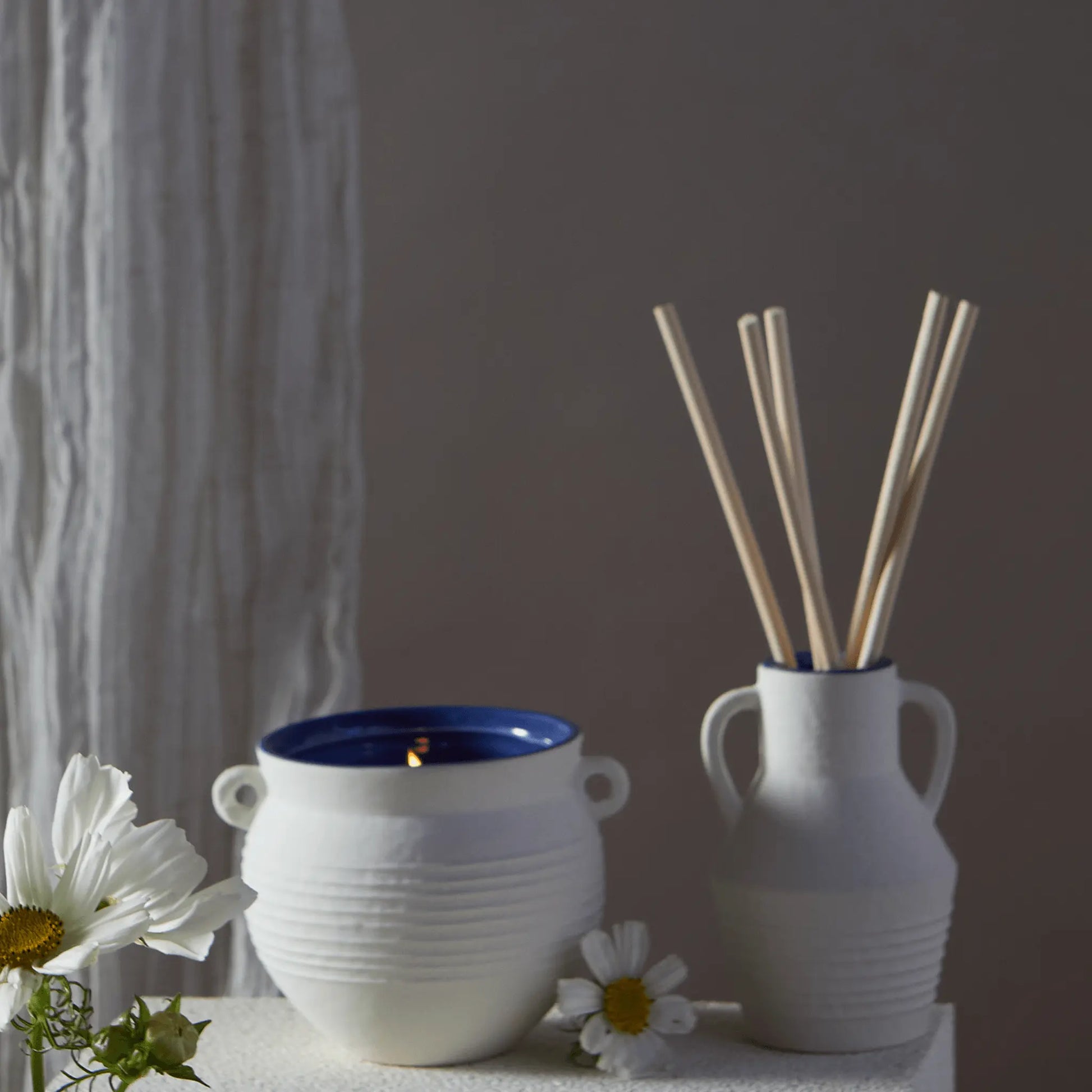 A white ceramic candle burns beside the Santorini 4 fl oz. Diffuser - Cliff & Hinoki Wood, which holds reed sticks and a daisy, evoking tranquil charm on a light surface.