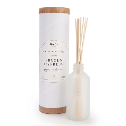 The Holiday Reed Diffuser - Frozen Cypress stands beside a frosted glass bottle with wooden reeds, providing a flameless fragrance ideal for holiday gifting.