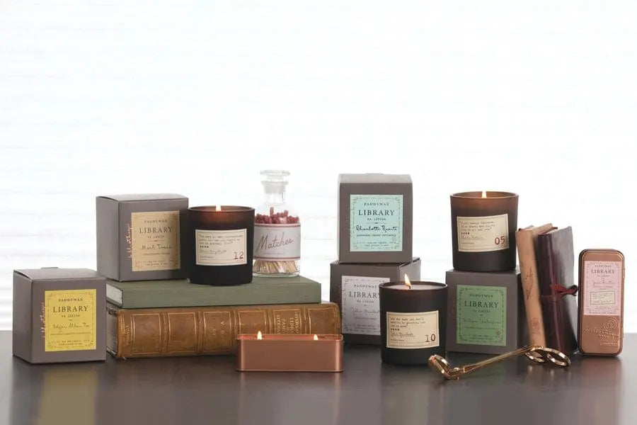 A collection of Paddywax Library candles in various scents, with vintage-inspired packaging and a few open books, a match jar, and a candle snuffer on a wooden surface.