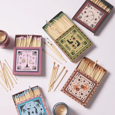 Four decorative matchboxes with astrological designs and colorful matchsticks are arranged on a light surface, surrounded by two small lit candles.