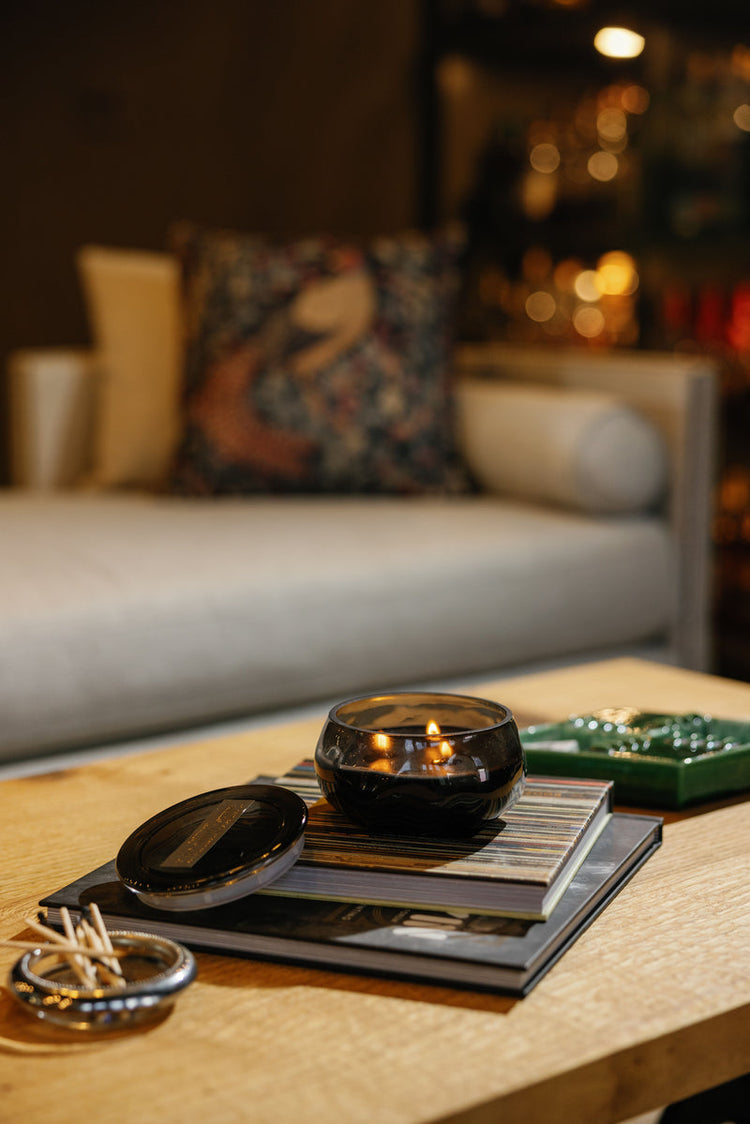 A black glass candle burns on a stack of books on a wooden coffee table. Matches and a decorative box sit nearby. A beige couch with a patterned pillow is in the background.