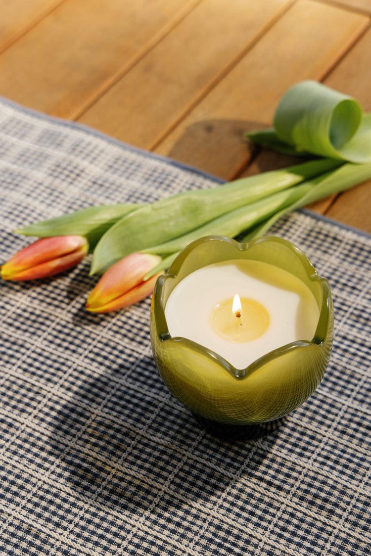 A lit candle in a green, flower-shaped holder sits on a checked cloth, with two pink tulips and green ribbon in the background on a wooden surface.