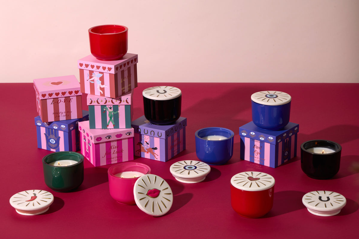 Several colorful candles with decorative lids are arranged on a pink and red surface, alongside patterned boxes in purple, pink, and blue tones.