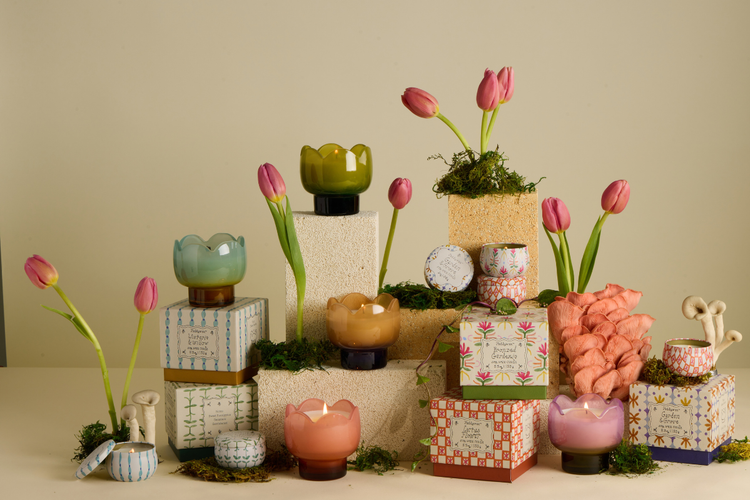 Colorful glass candle holders and decorative boxes are arranged on textured blocks with pink tulips and moss, creating a whimsical, floral-themed display against a neutral background.