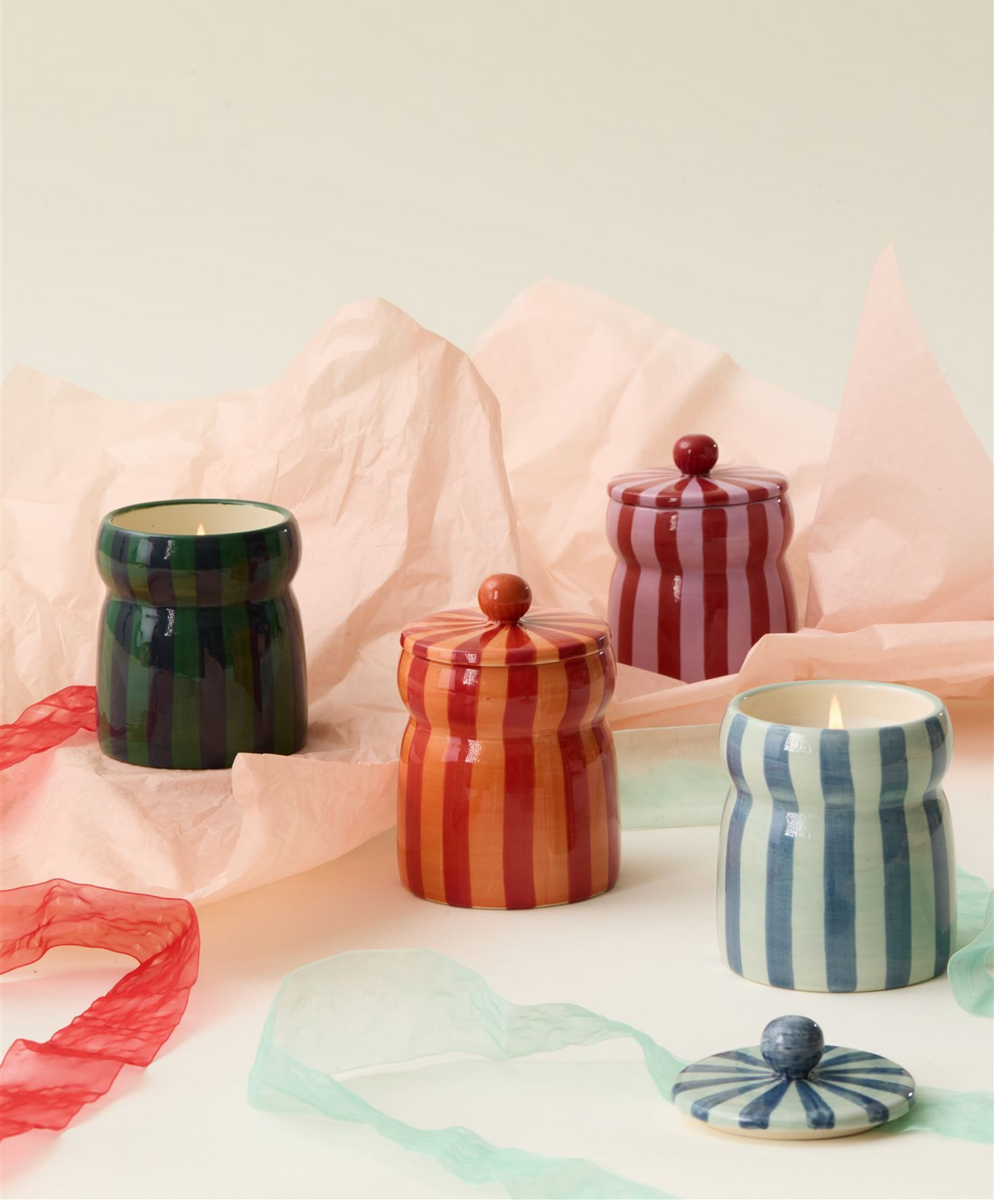Four striped ceramic candles in green, red, orange, and blue, some with lids on, are arranged on soft pink tissue paper with colorful ribbons on a light background.