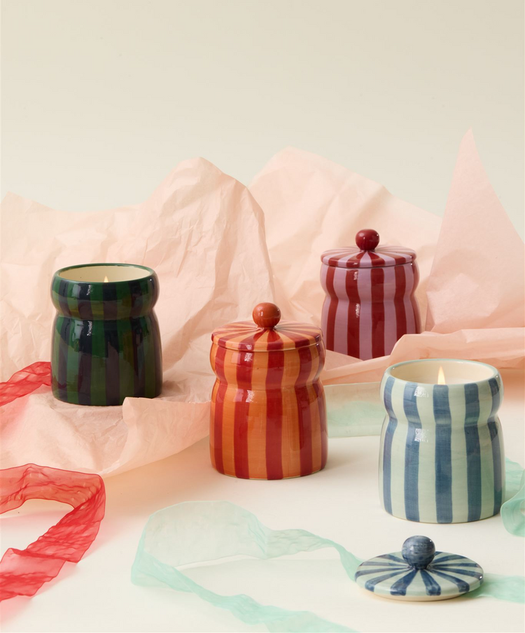 Four striped ceramic candles in green, red, orange, and blue, some with lids on, are arranged on soft pink tissue paper with colorful ribbons on a light background.