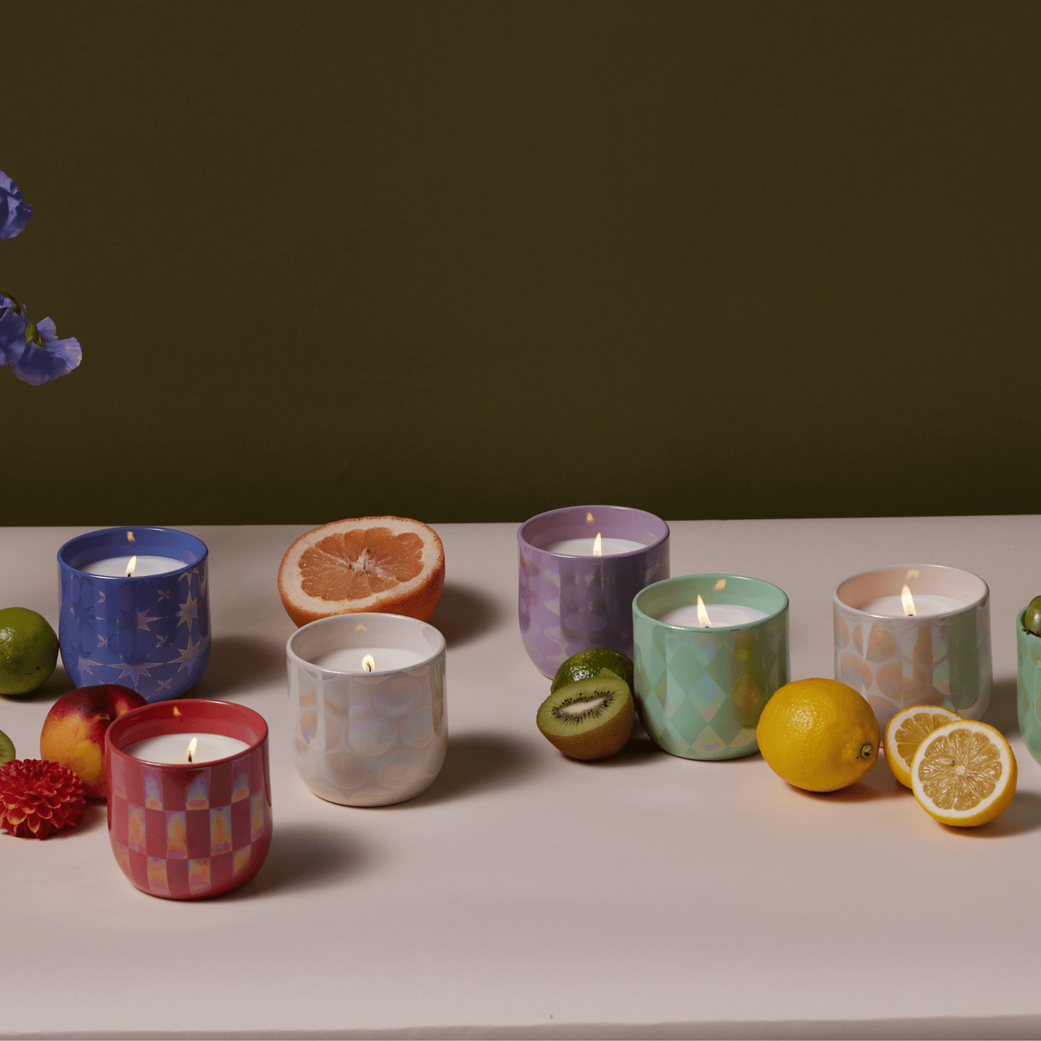 Lustre collection candles on a pale surface with fruit and a dark backdrop