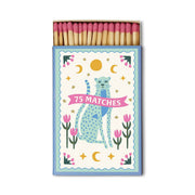 A Dopo “Blue Cheetah” - 75ct Boxed Matches are perfect for candle lovers, showcasing a blue illustrated cat surrounded by pink tulips and gold stars. The box is labeled “75 MATCHES” and contains wooden safety matches with pink tips.