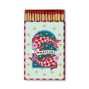 A Dopo candle lovers will appreciate the A Dopo “Koi” - 75ct Boxed Matches, featuring a colorful design of two koi fish, gold stars, and the words “75 Matches” on the front. The open box displays safety matches with red tips inside.