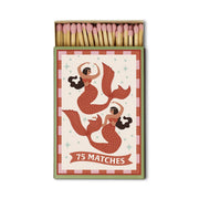 A Dopo “Mermaid & Moon” boxed matches contain 75 safety matches in a box adorned with two illustrated mermaids featuring red tails and brown hair, set against a decorative background inspired by the allure of Mediterranean travel.