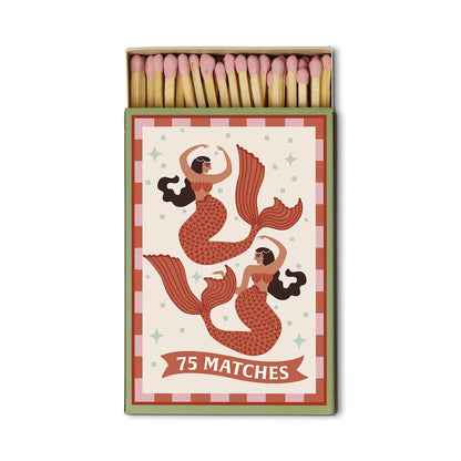 A Dopo “Mermaid & Moon” boxed matches contain 75 safety matches in a box adorned with two illustrated mermaids featuring red tails and brown hair, set against a decorative background inspired by the allure of Mediterranean travel.
