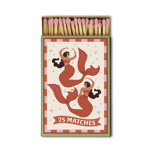 A Dopo “Mermaid & Moon” boxed matches contain 75 safety matches in a box adorned with two illustrated mermaids featuring red tails and brown hair, set against a decorative background inspired by the allure of Mediterranean travel.