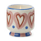 The A Dopo Hearts Candle - Rosewood Vanilla features a lit ceramic holder with pink hearts, blue dots, and stripes on white, delivering bold Mediterranean-inspired scents.