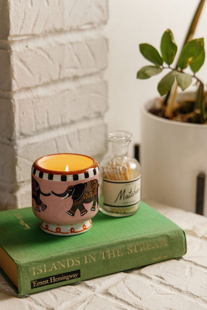 A lit Tobacco & Vanilla - A Dopo 8 oz Candle rests on a green copy of "Islands in the Stream" by Ernest Hemingway, beside a glass match jar and a potted plant.