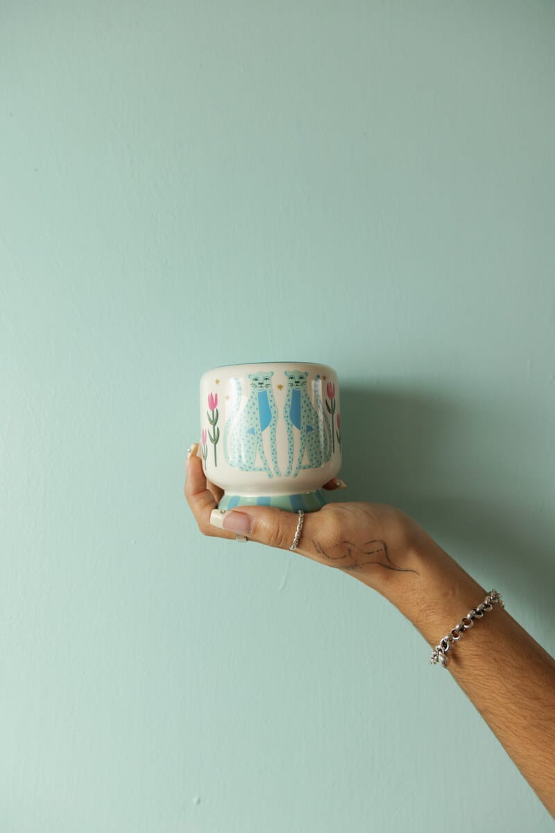 A hand adorned with silver rings and a bracelet holds the Linen & Orris - A Dopo 8 oz Candle, a small, round, hand-painted ceramic cup featuring blue cats and pink flowers, set against a light blue wall.