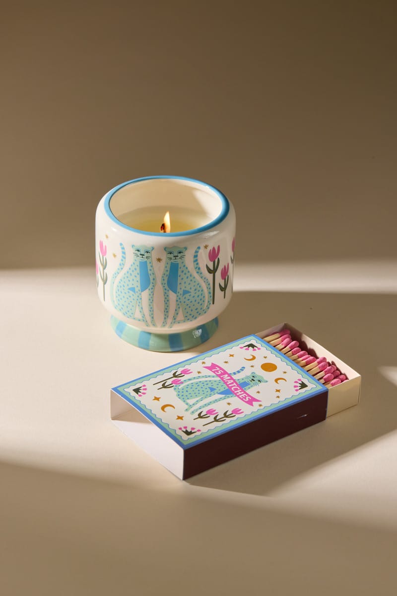 A lit A Dopo candle in a decorative holder with a cat and floral design sits beside an open box of A Dopo “Blue Cheetah” - 75ct Boxed Matches, also adorned with a similar cat illustration. Soft shadows fall on the beige surface.