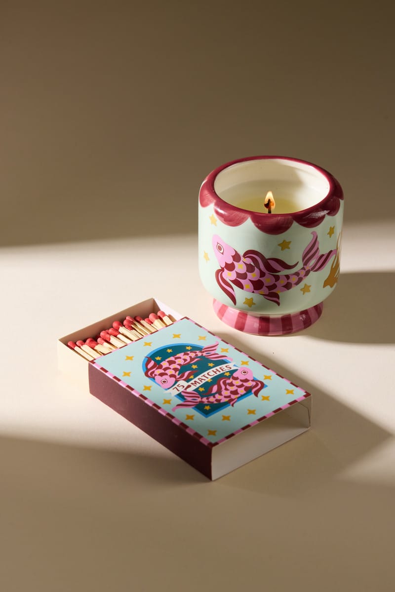 A lit A Dopo candle in a decorative fish-themed holder sits beside an open box of A Dopo “Koi” 75ct matches, featuring pink-tipped safety matches on a beige surface illuminated by soft light.