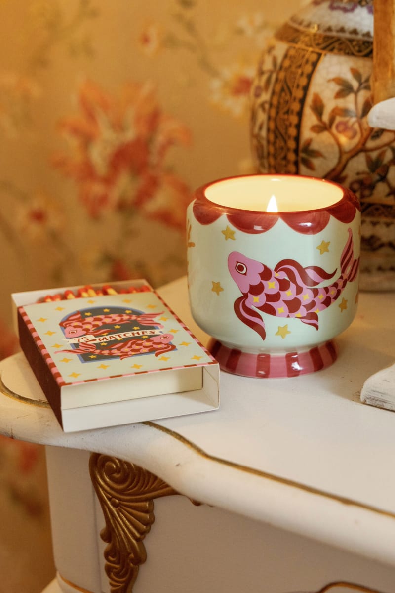 An Ocean Tide - A Dopo 8 oz Candle, lit and presented in a decorative fish-patterned holder, rests on a white table next to an open box of matches, with floral wallpaper and part of a patterned vase visible in the background.