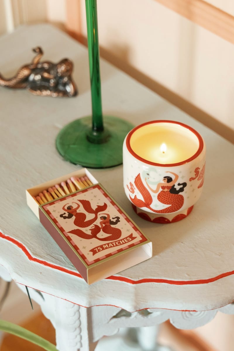 An A Dopo Ginger & Teak 8 oz candle, featuring a red dragon design, sits beside a box of matches on a light blue table near a green candlestick and a small metallic decorative object, filling the space with its warm, earthy scent.