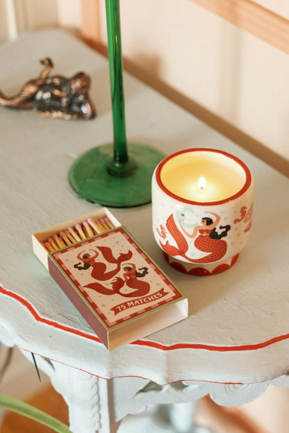 An A Dopo Ginger & Teak 8 oz candle, featuring a red dragon design, sits beside a box of matches on a light blue table near a green candlestick and a small metallic decorative object, filling the space with its warm, earthy scent.