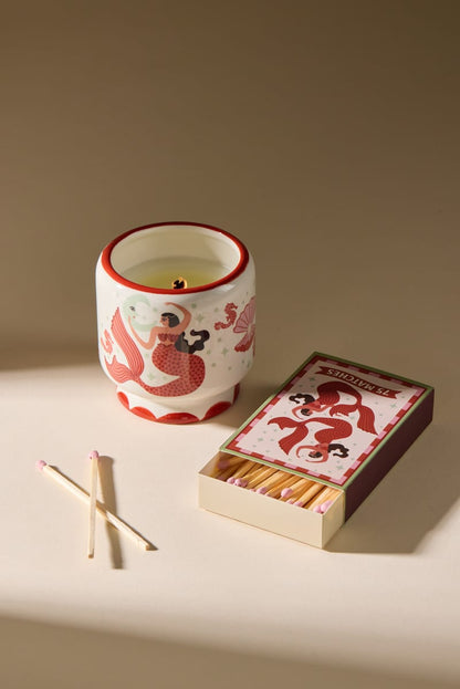 A white candle holder adorned with red designs of women and fish sits beside the A Dopo “Mermaid & Moon” - 75ct Boxed Matches, with three matches lying on a beige surface. Warm light casts soft shadows, creating a cozy, inviting atmosphere reminiscent of Mediterranean travel.
