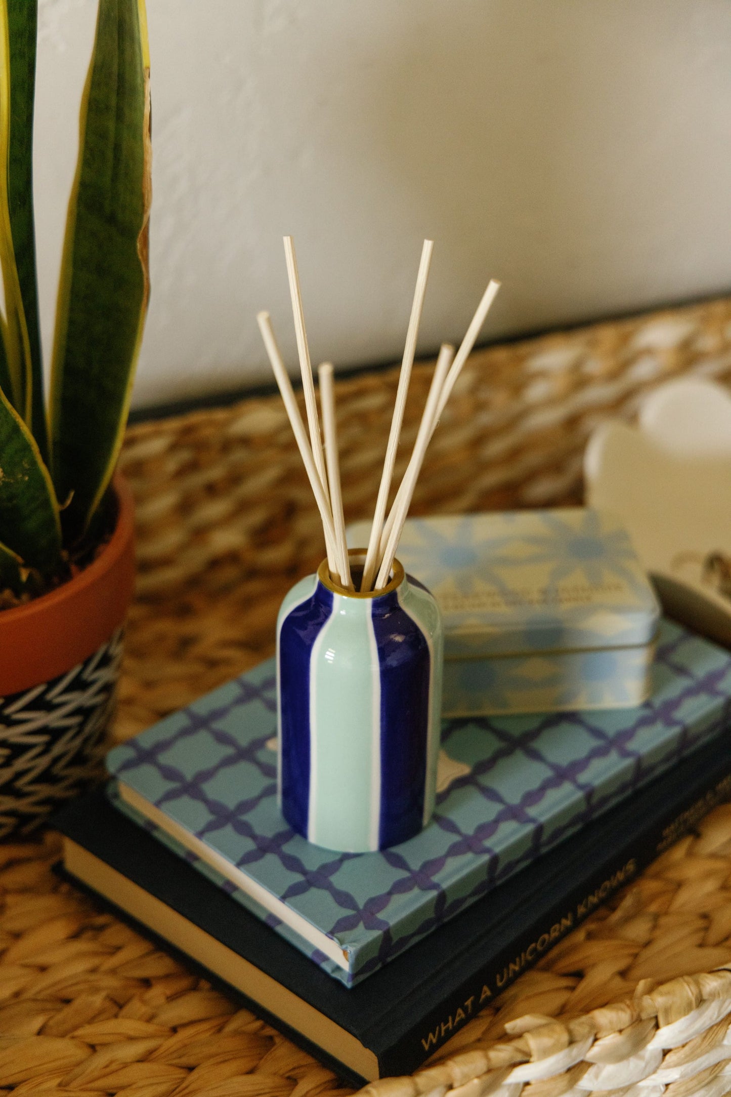 The Incense & Smoke - A Dopo 4 oz Diffuser, featuring blue and green stripes and White Grapefruit fragrance, is displayed with reed sticks on decorative books and a tin beside a potted plant on a woven tray.