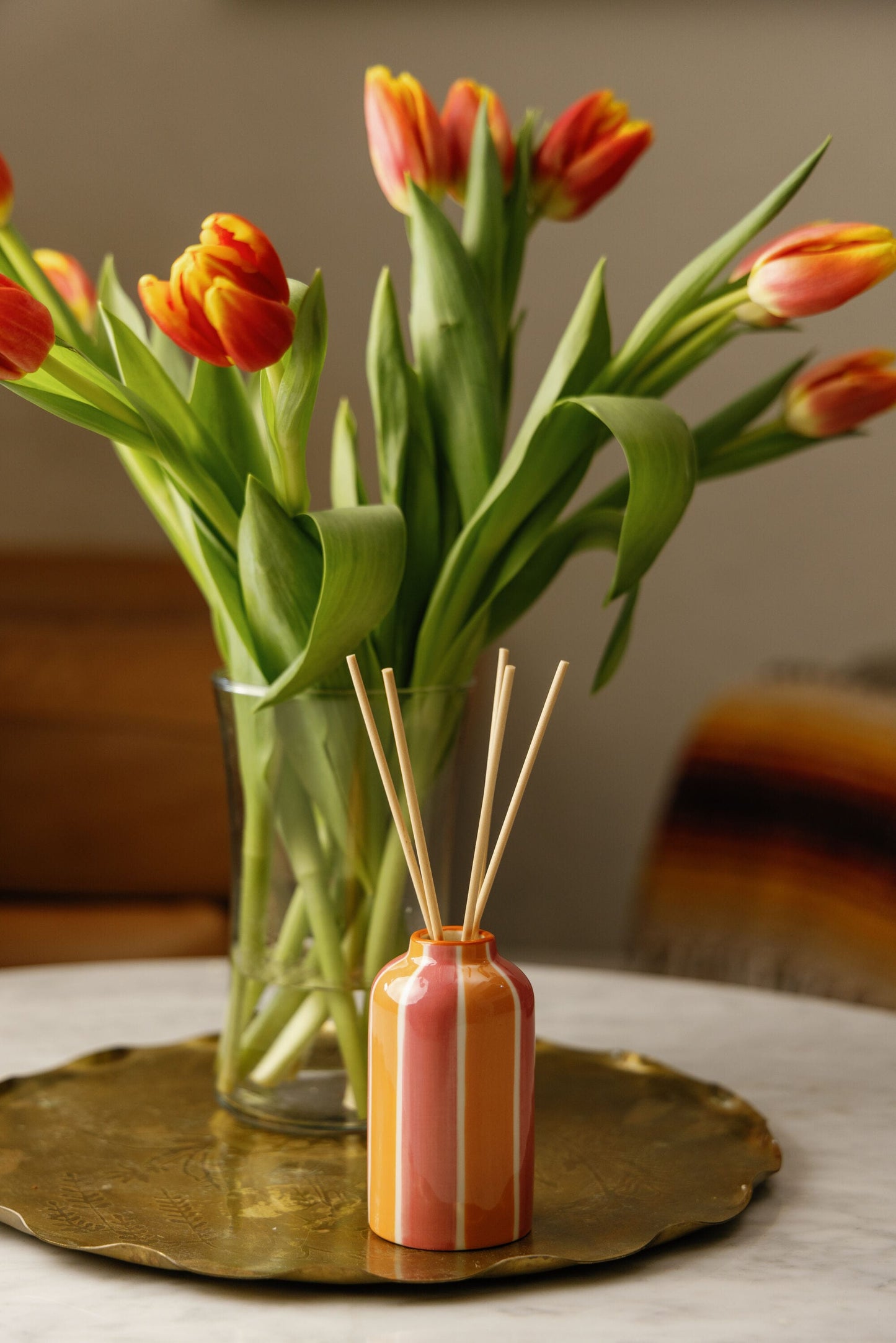 A glass vase with orange and yellow tulips sits on a round brass tray next to the Orange Blossom - A Dopo 4 oz Diffuser, displayed on a marble surface with a softly blurred background.