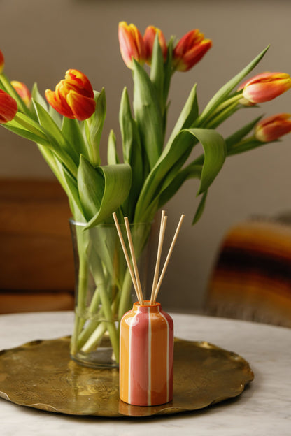 A glass vase with orange and yellow tulips sits on a round brass tray next to the Orange Blossom - A Dopo 4 oz Diffuser, displayed on a marble surface with a softly blurred background.