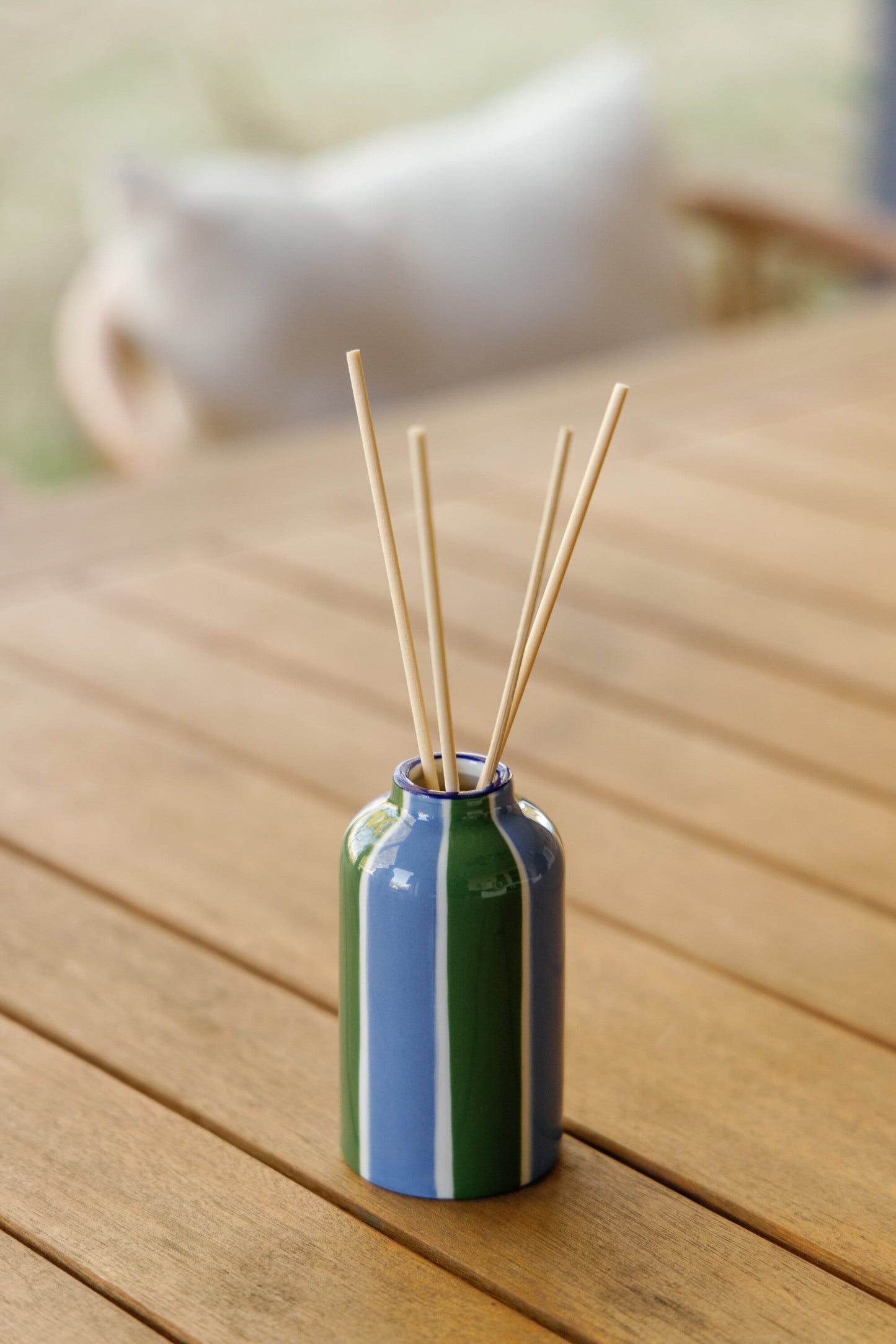 The Misted Lime - A Dopo 4 oz Diffuser features a small ceramic vase with blue and green vertical stripes, holding thin wooden reeds, displayed on a wooden table with a blurred background and cushion visible.