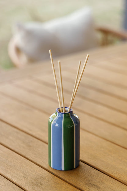 The Misted Lime - A Dopo 4 oz Diffuser features a small ceramic vase with blue and green vertical stripes, holding thin wooden reeds, displayed on a wooden table with a blurred background and cushion visible.