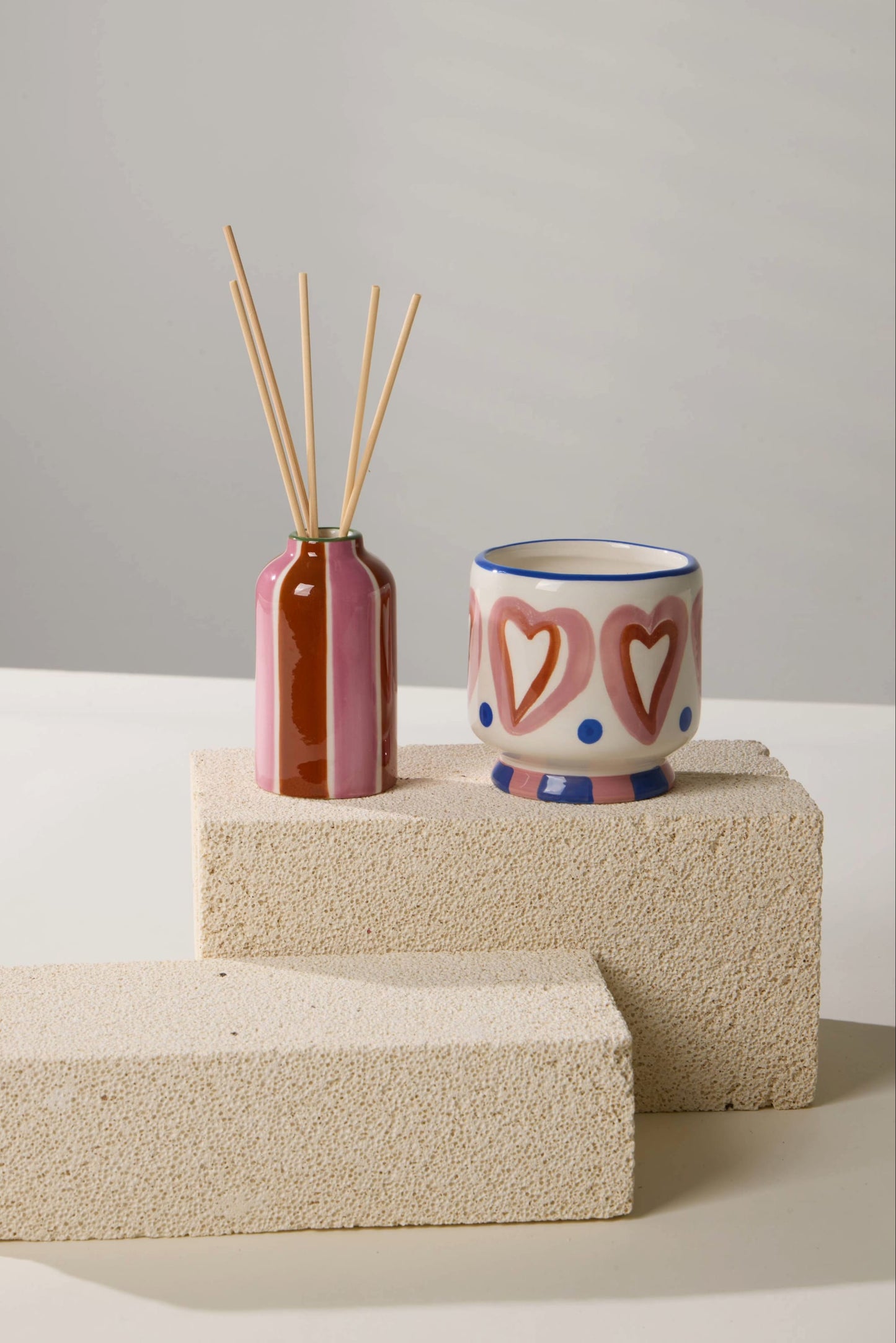 A Black Cedar & Fig - A Dopo 4 oz Diffuser and a ceramic cup with pink hearts and blue dots sit on two stacked, textured beige blocks against a neutral background.