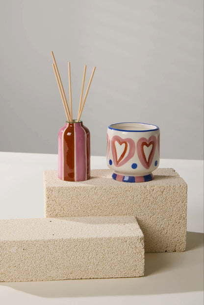 A Black Cedar & Fig - A Dopo 4 oz Diffuser and a ceramic cup with pink hearts and blue dots sit on two stacked, textured beige blocks against a neutral background.